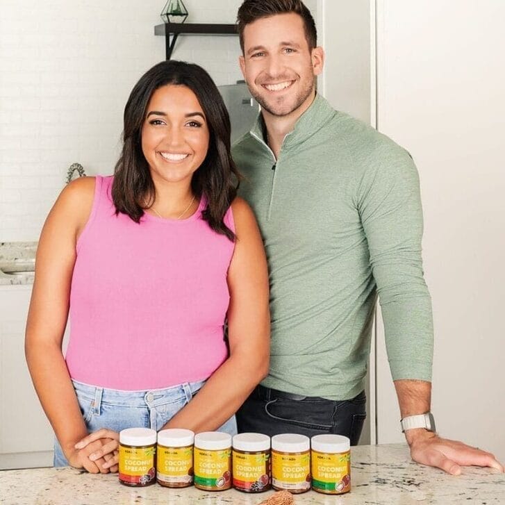 A woman in a pink shirt and a man in a light green shirt stand together behind a marble countertop that has six jars of coconut spread on it.