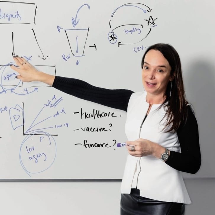 Professor Cait Lamberton in a white shirt with black sleeves gestures at handwritten notes on a whiteboard.