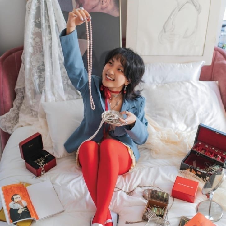 A woman in a blue suit jacket, red leggings, and silver shoes sits on a bed with books and jewelry around her and holds up a pearl necklace.
