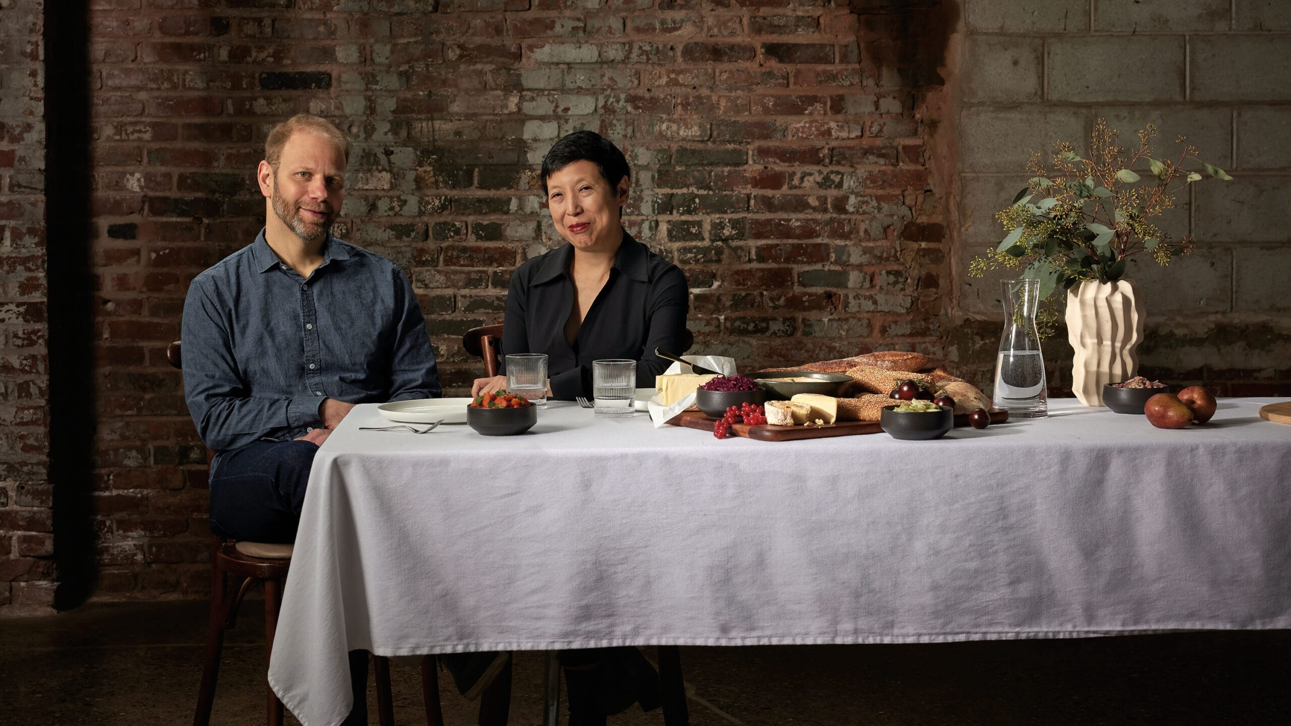 A man in a denim button-down shirt and a woman in a black button-down shirt sit together against a brick wall in front of a table covered with a white tablecloth, glasses and a pitcher of water, a vase, bread, cheese, and fruits.