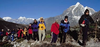 About a dozen people hike in front of a snow-capped mountain.