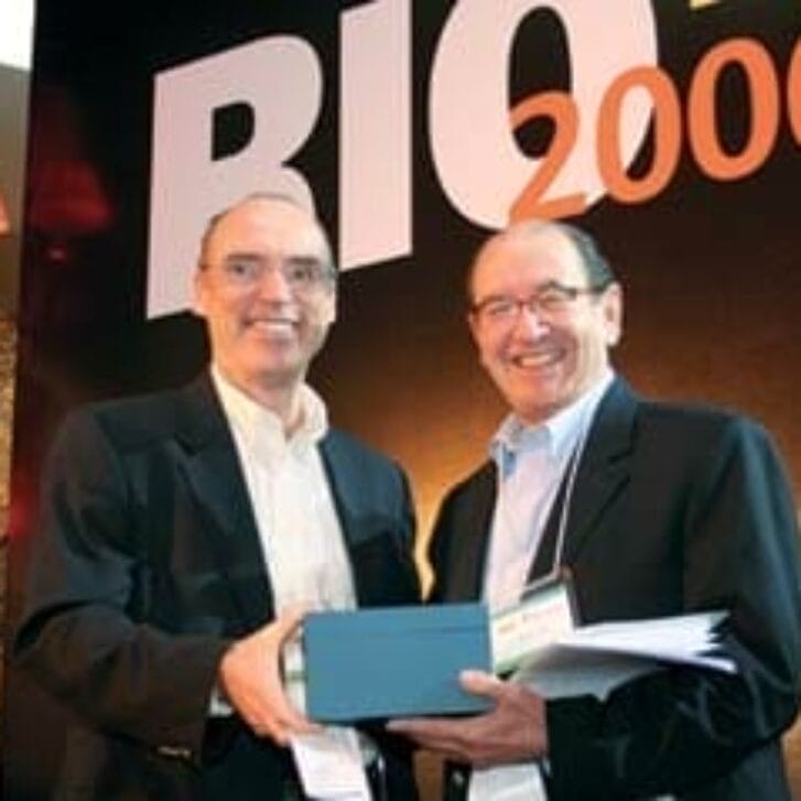Two men in business attire stand together for a photo in front of a sign with Rio 2006 written on it.
