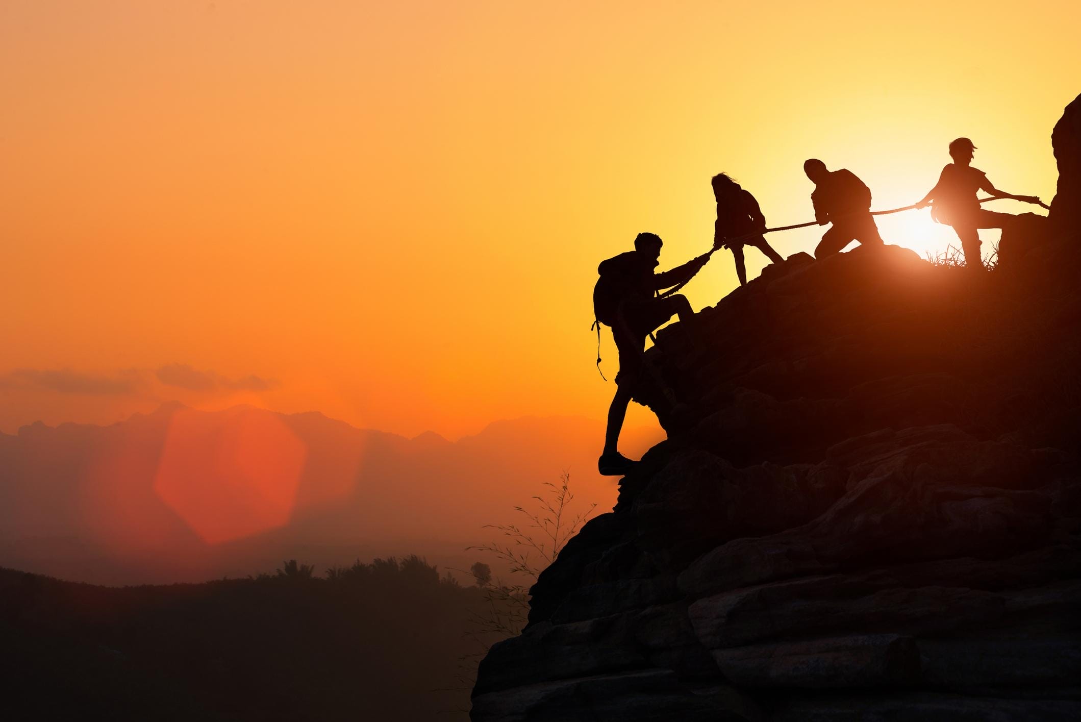 Four rock climbers help each other scale a mountain at sunset.