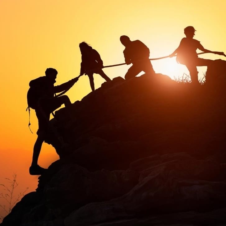 Four rock climbers help each other scale a mountain at sunset.