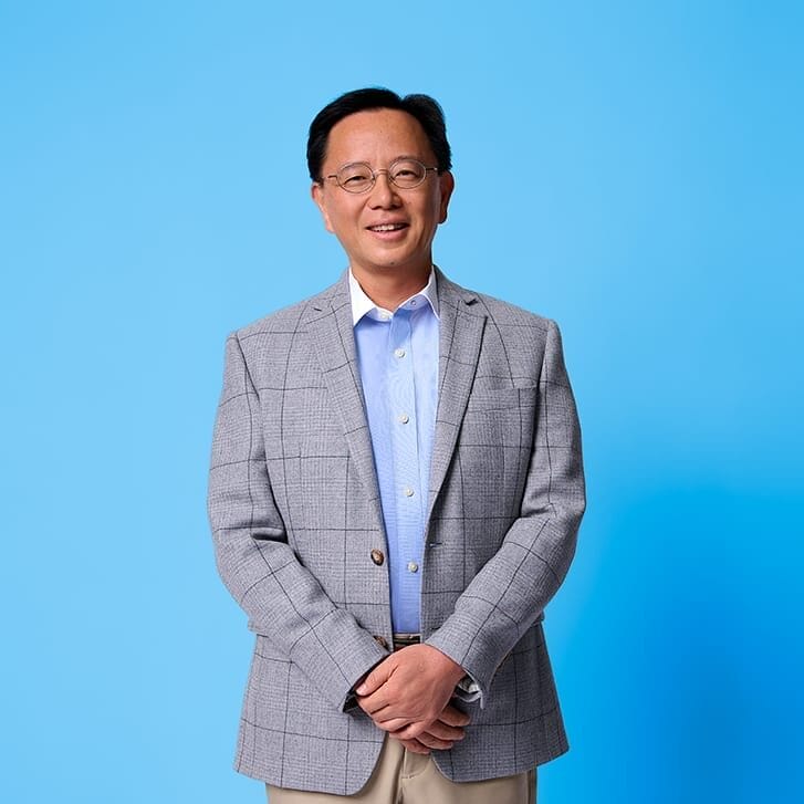 A man in a gray blazer, light blue shirt, and tan khakis poses for a photo in front of a light blue background.