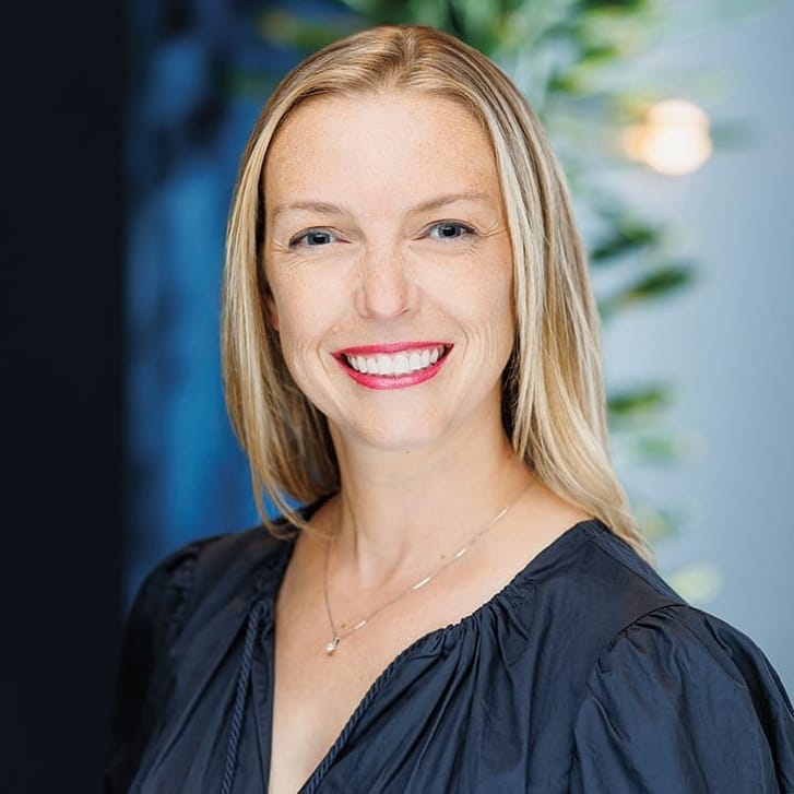 Portrait of a woman with blond hair wearing a silver necklace and a semi-formal blue shirt.