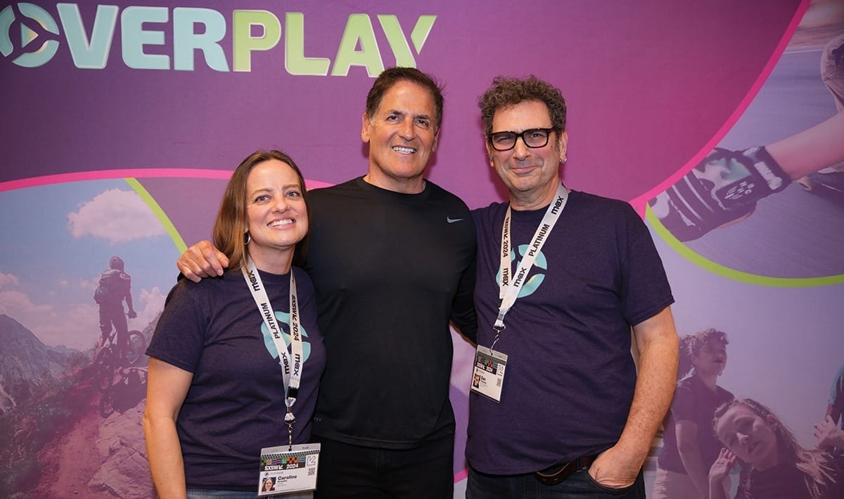 Three people pose for a photo together against a backdrop with imagery for the startup Overplay on it.