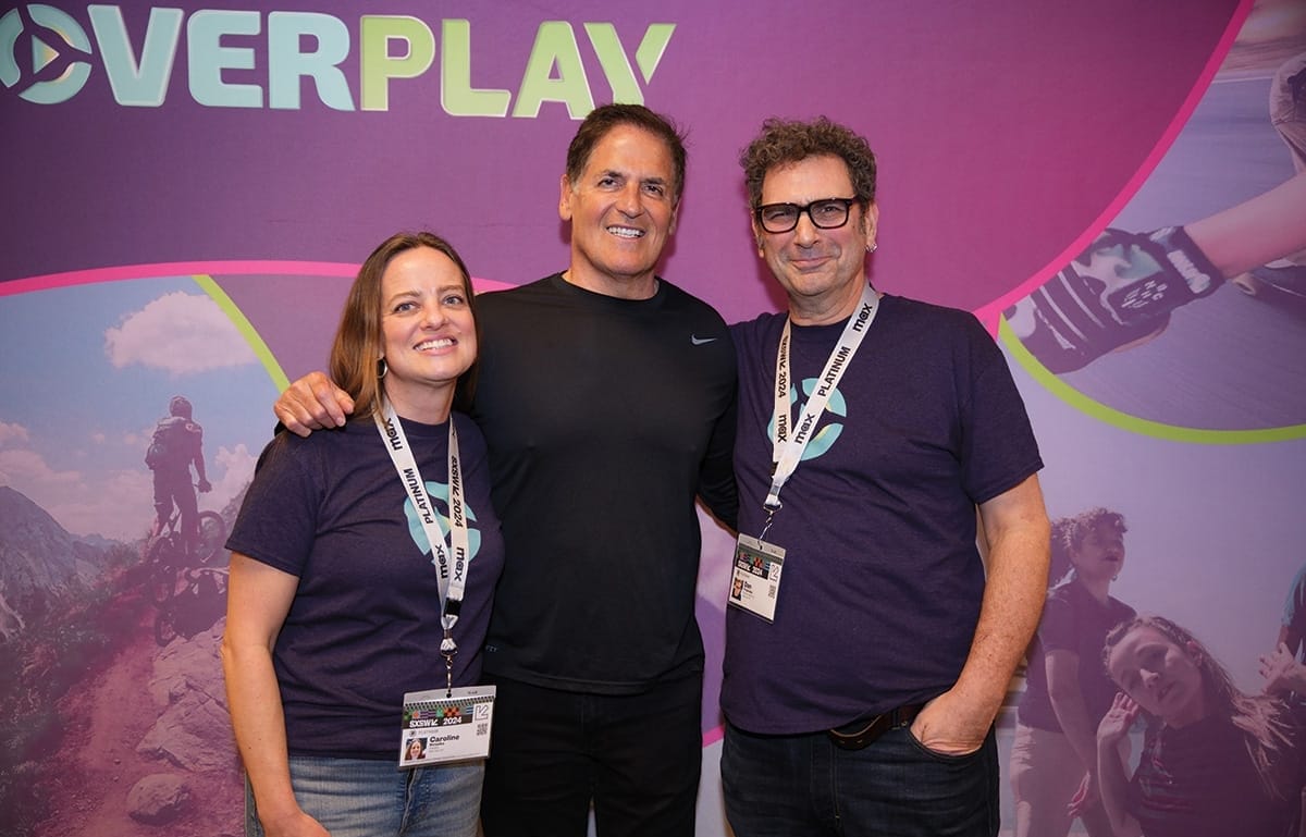 Three people pose for a photo together against a backdrop with imagery for the startup Overplay on it.