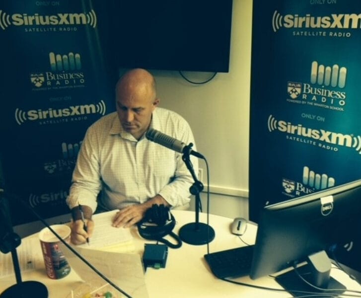 Radio Host Dan Loney writes notes while seated at a table in front of a microphone.