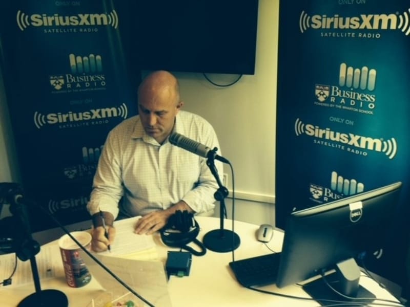 Radio Host Dan Loney writes notes while seated at a table in front of a microphone.