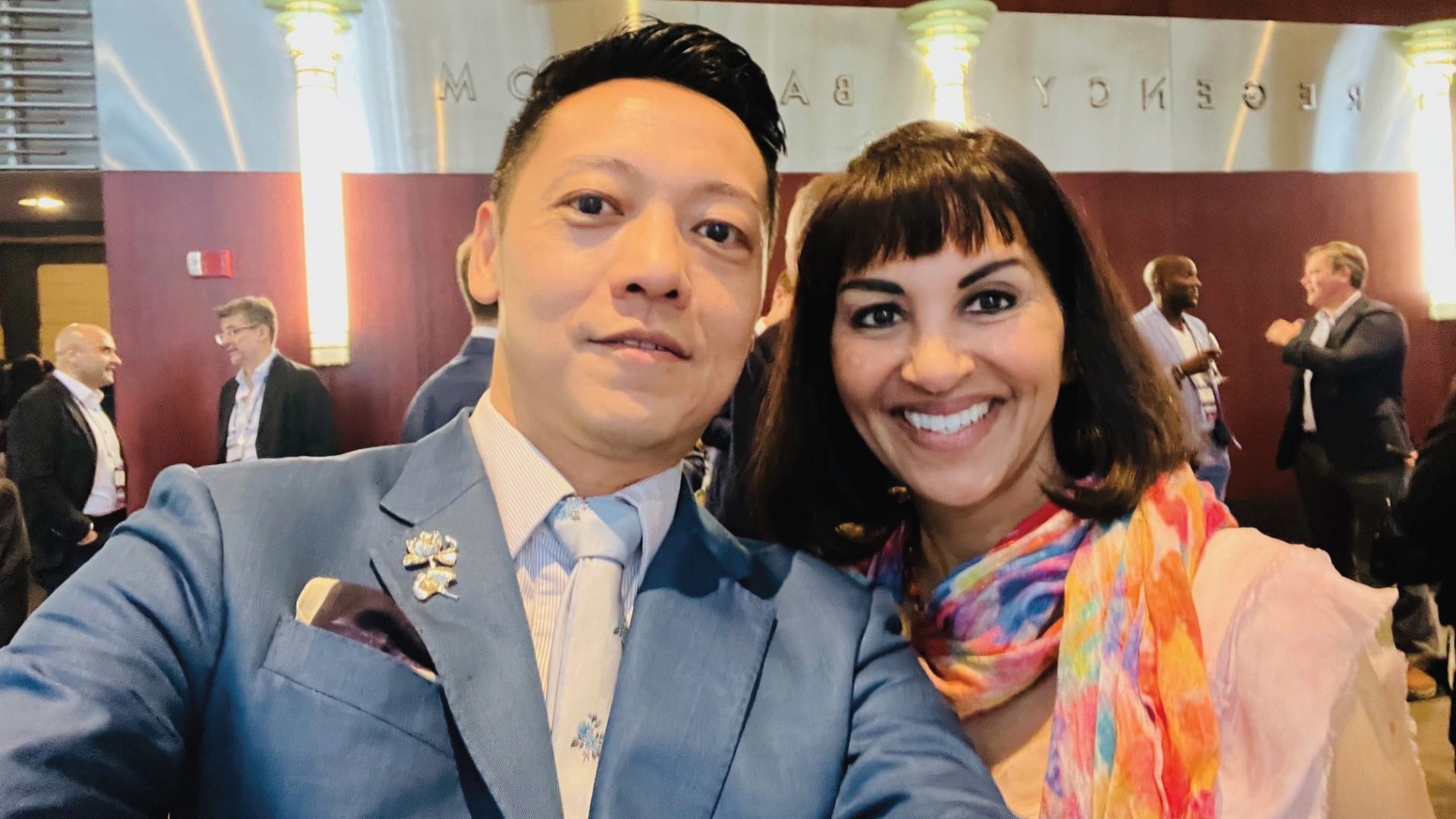 A man in a light blue suit and a woman in colorful attire pose for a selfie.