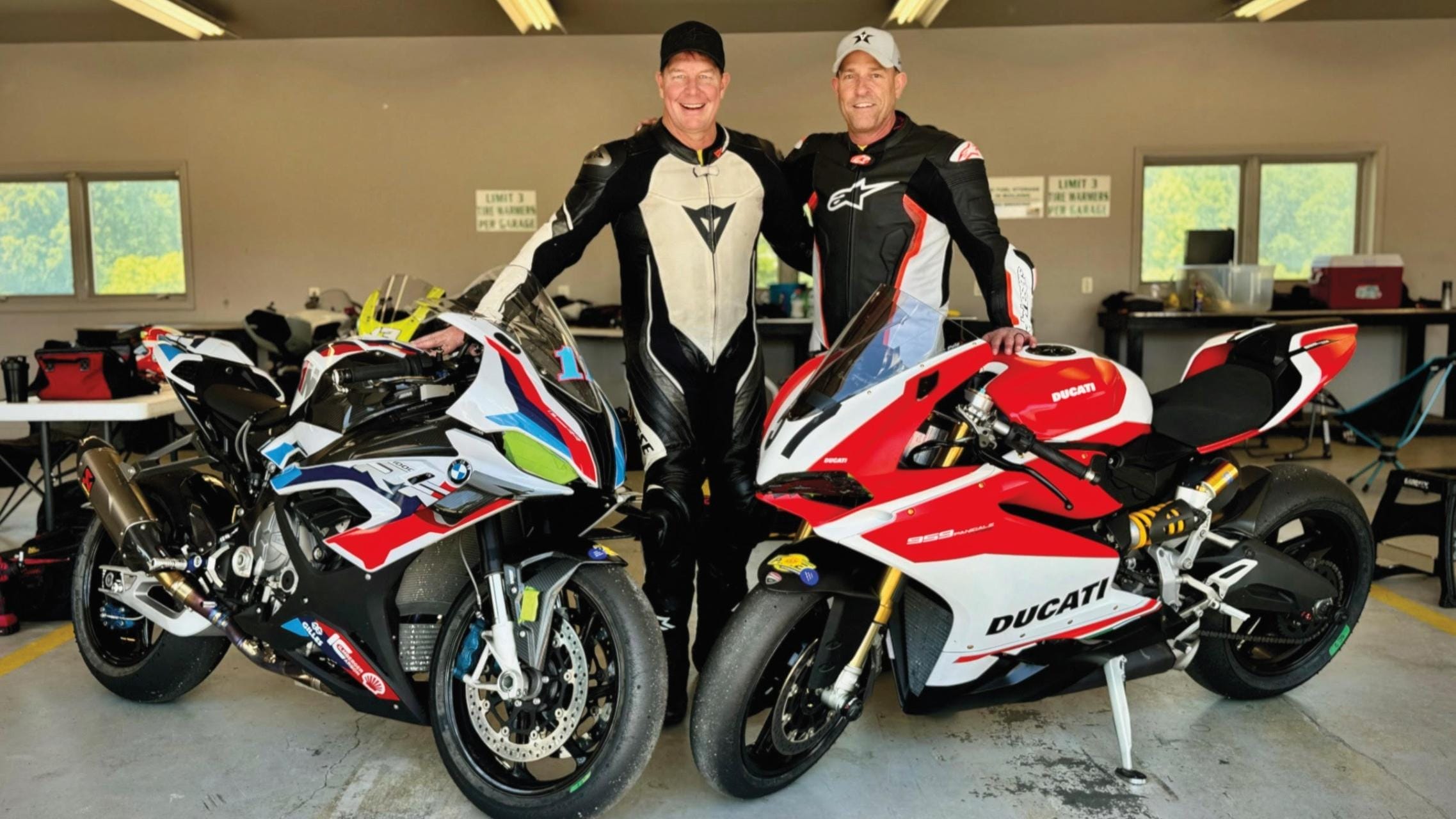 Two men in motorcycle suits pose in front of two motorcycles.