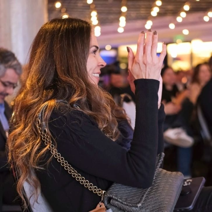 A woman in a black shirt seated in an audience claps her hands.