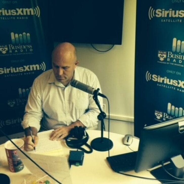 Radio Host Dan Loney writes notes while seated at a table in front of a microphone.