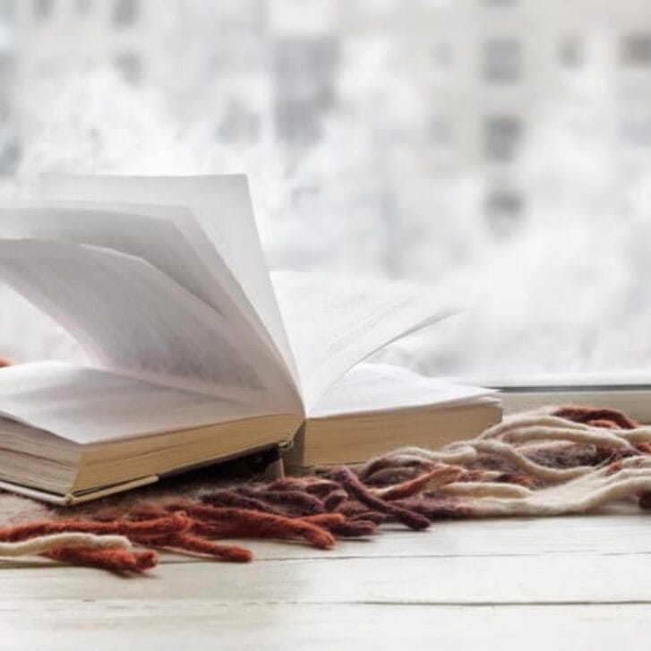 An open book next to a window with snow falling outside.