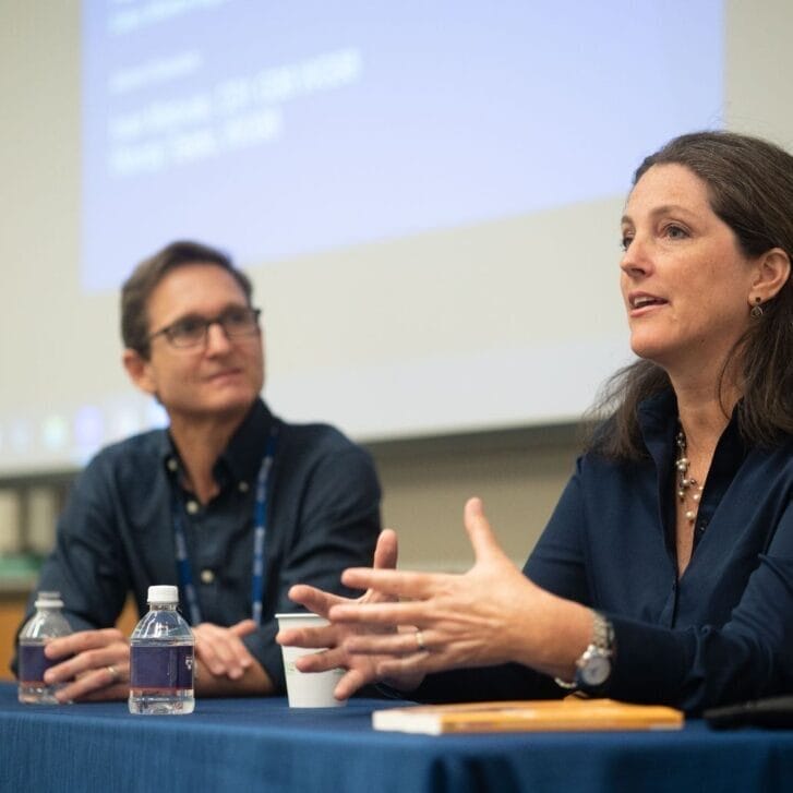 A man and woman both wearing blue semi-formal clothing sit at a table in a classroom, the woman gesturing as she speaks.