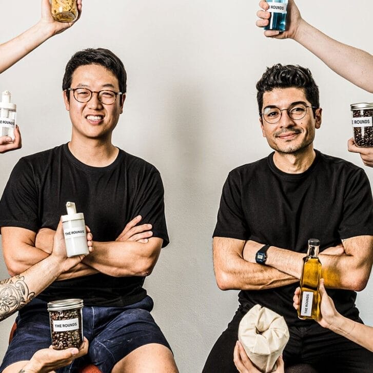 Two men in black shirts sit on stools with hands reaching into the frame with household goods in hand.