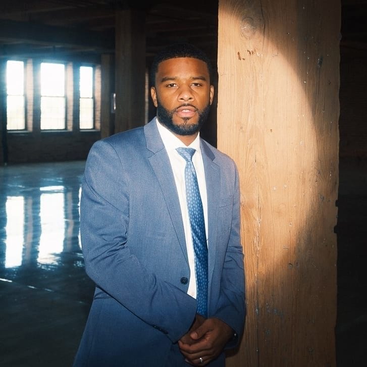 Portrait of a suited-up Ross Mac stands in a darkly lit room with some light streaming through a set of windows and a spotlight on the subject.