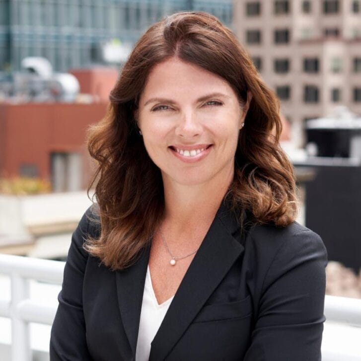 Woman in a black blazer and white shirt poses against a cityscape.