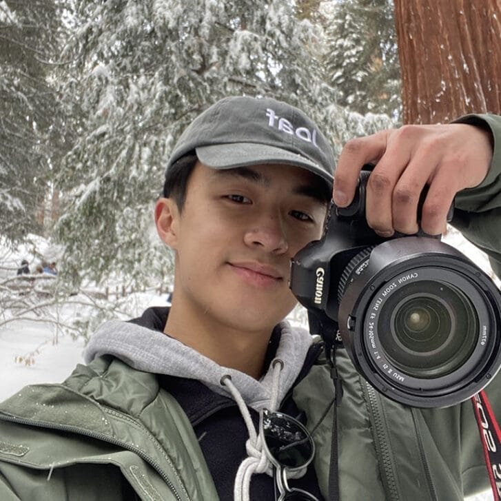 Self-portrait of Jesse Zhang holding a camera among redwood trees in the snow.