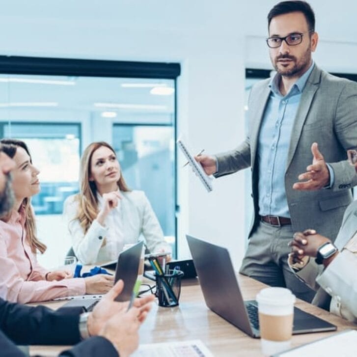 Man standing in a board room, gesturing widely in front of colleagues