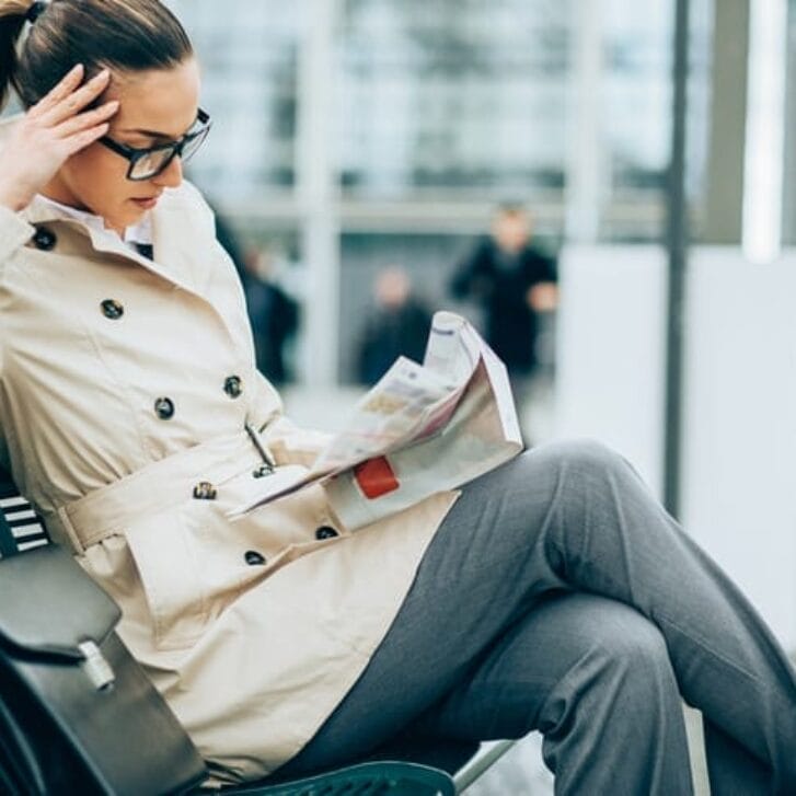 Young woman in business wear sitting on a bench in modern urban setting and reading a newspaper.