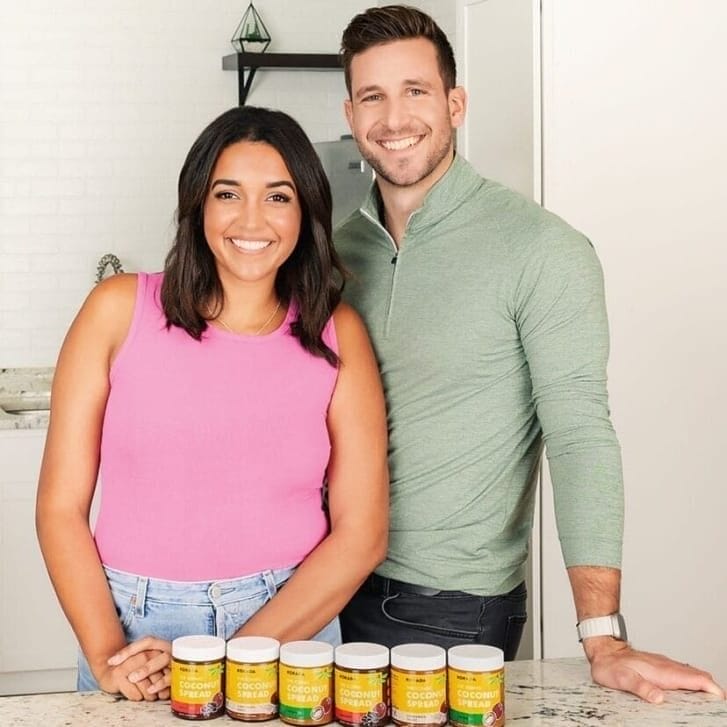 A woman in a pink shirt and a man in a light green shirt stand together behind a marble countertop that has six jars of coconut spread on it.