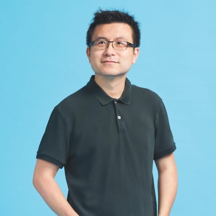 A man in a black polo shirt, pants, and glasses standing in front of a light blue backdrop.