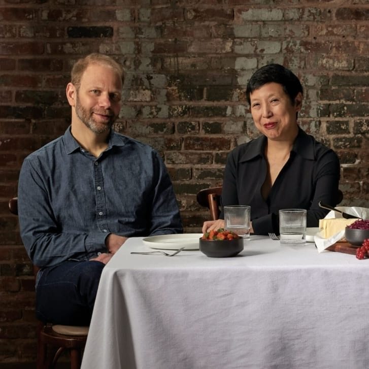 A man in a denim button-down shirt and a woman in a black button-down shirt sit together against a brick wall in front of a table covered with a white tablecloth, glasses and a pitcher of water, a vase, bread, cheese, and fruits.