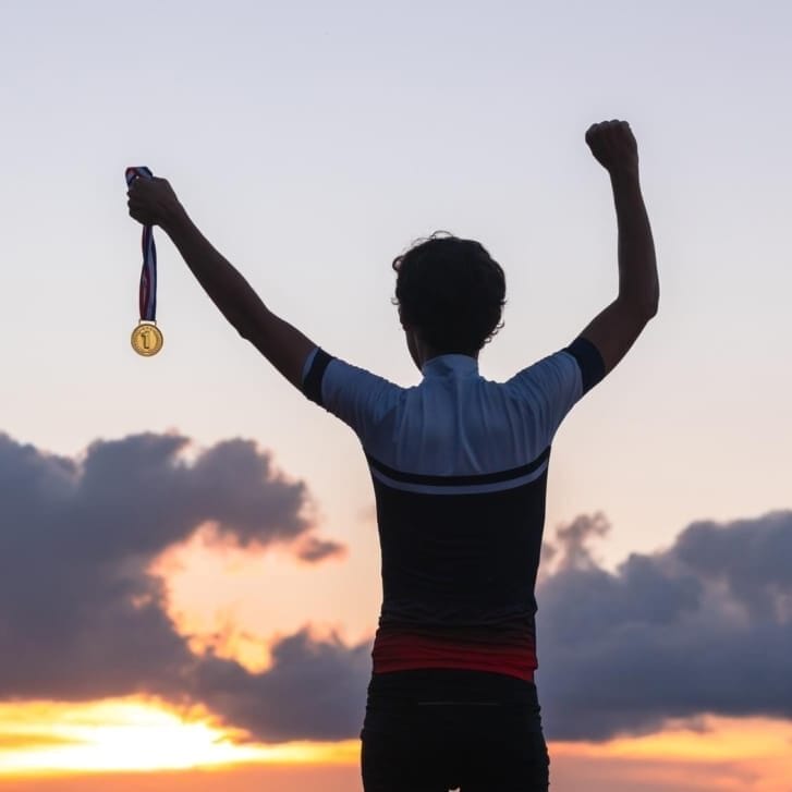 Man's silhouette as he holds a gold medal up and stands in front of a sunrise.