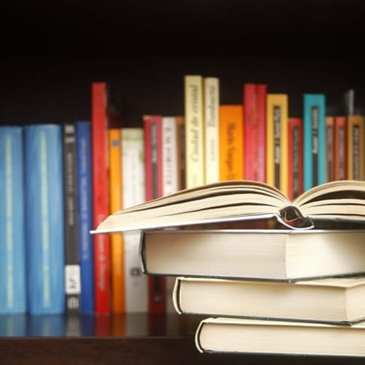 Stack of books on a wooden library shelf, the one on top open , and multicolored book spines in a row in the background.