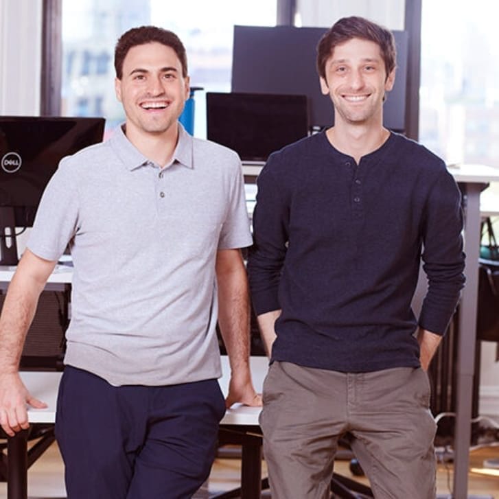 Two men stand side-by-side posing for a photo leaned against a desk, with computer monitors behind them.