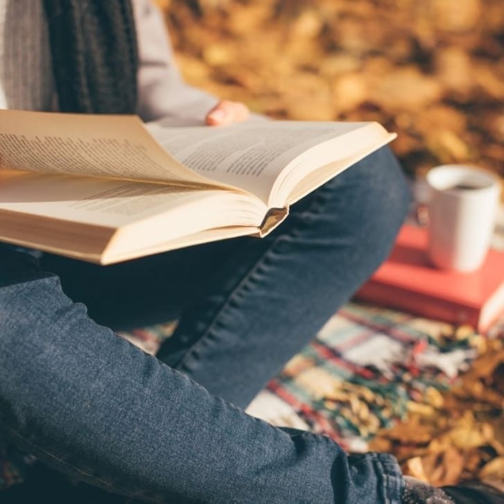 A woman sitting on a blanket, reading a book, and drinking coffee in an autumn garden.