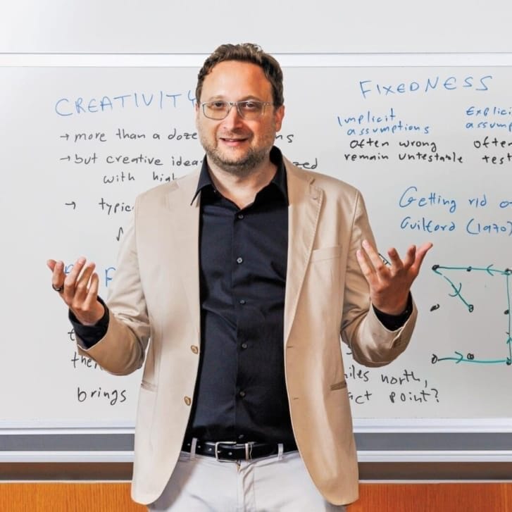 A man in a tan suit jacket, black shirt, and white pants stands and gestures in front of a whiteboard filled with written notes.