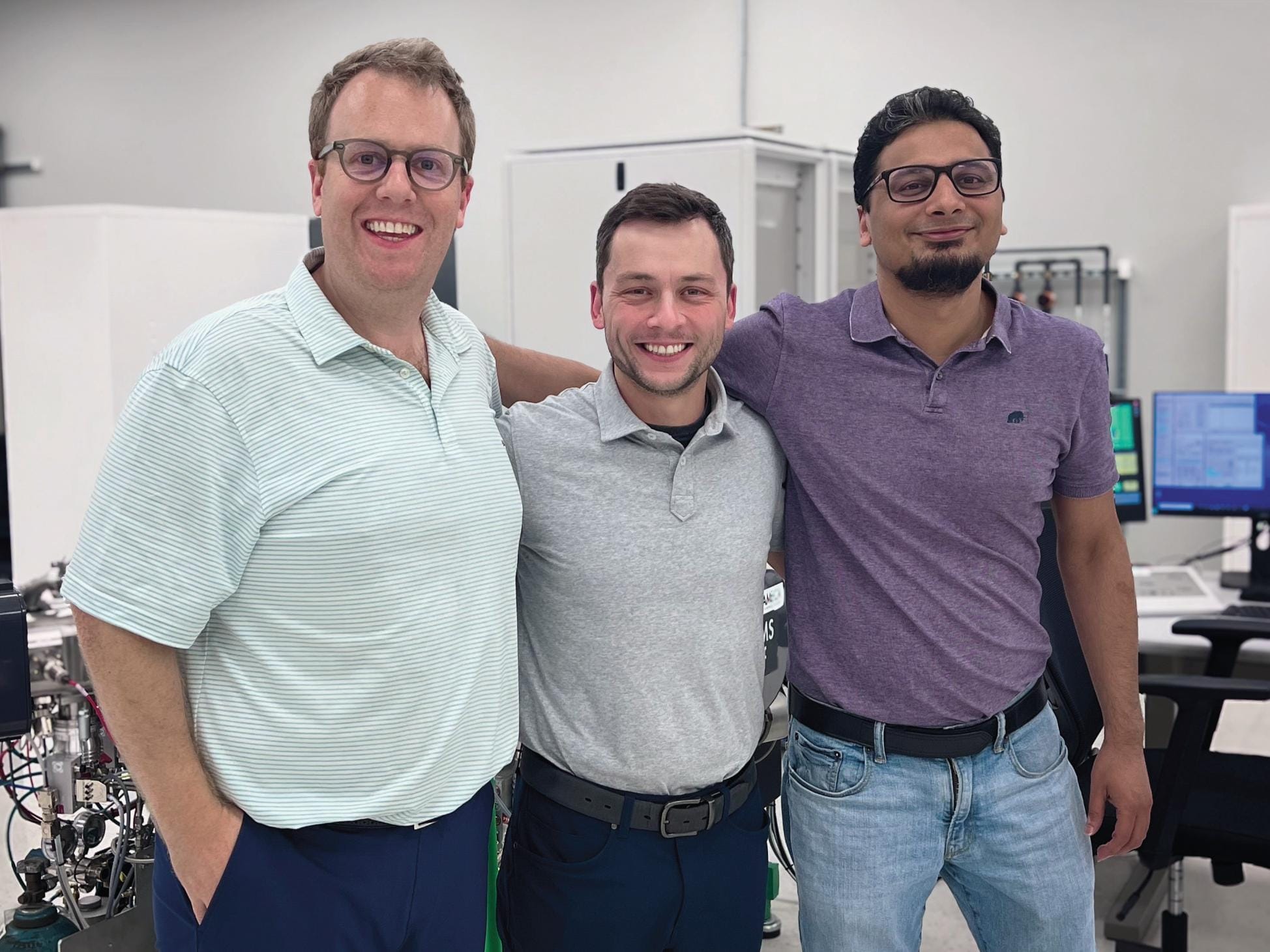 Three men stand shoulder to shoulder wearing polos in different colors in a lab-like setting.