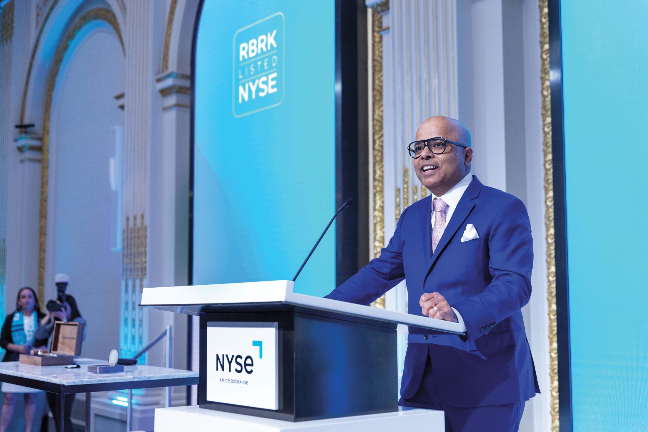 A man in a blue sit stands at a podium at the New York Stock Exchange.