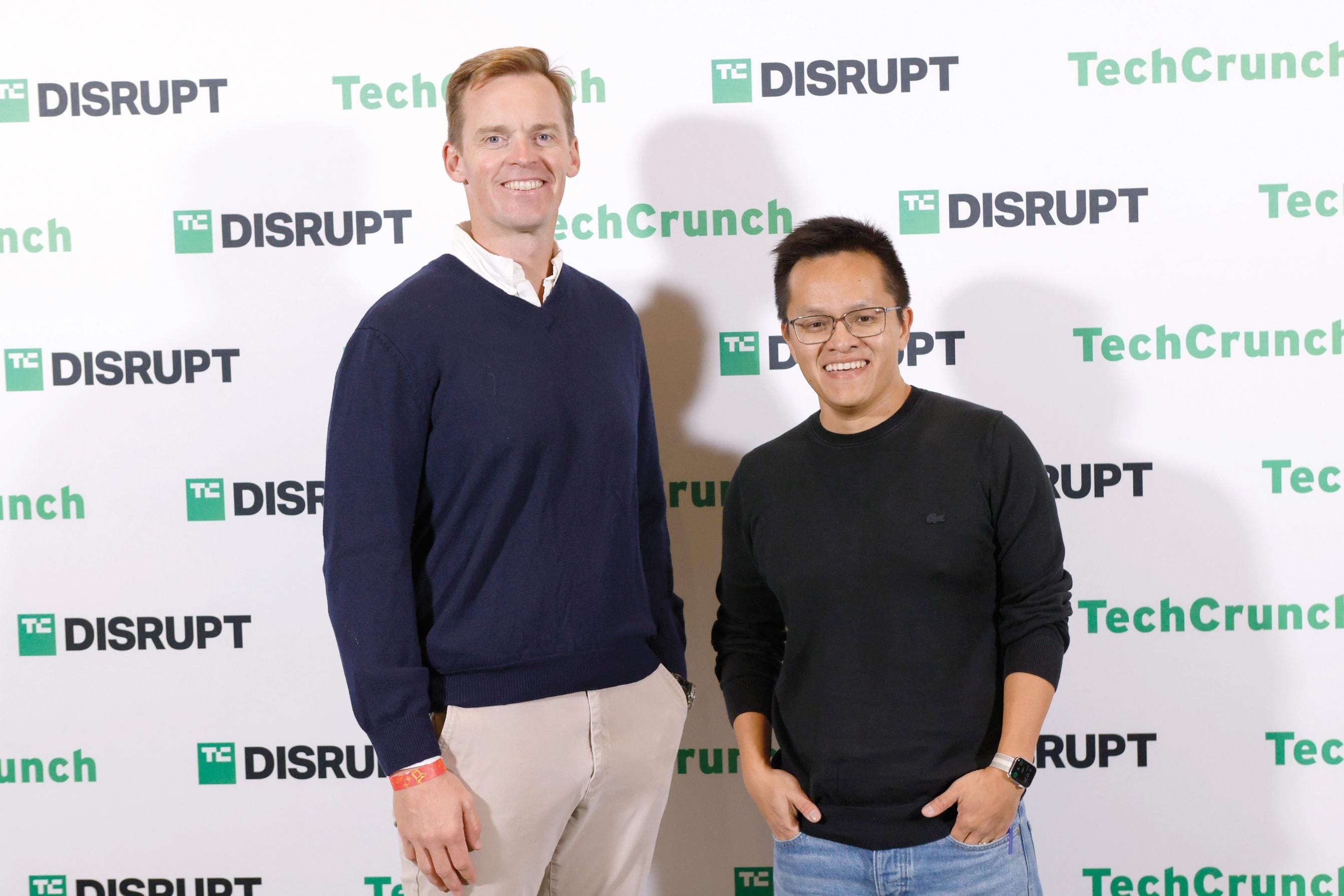 Two men stand in long sleeve shirts posed in front of a photo background that says TechCrunch Disrupt.