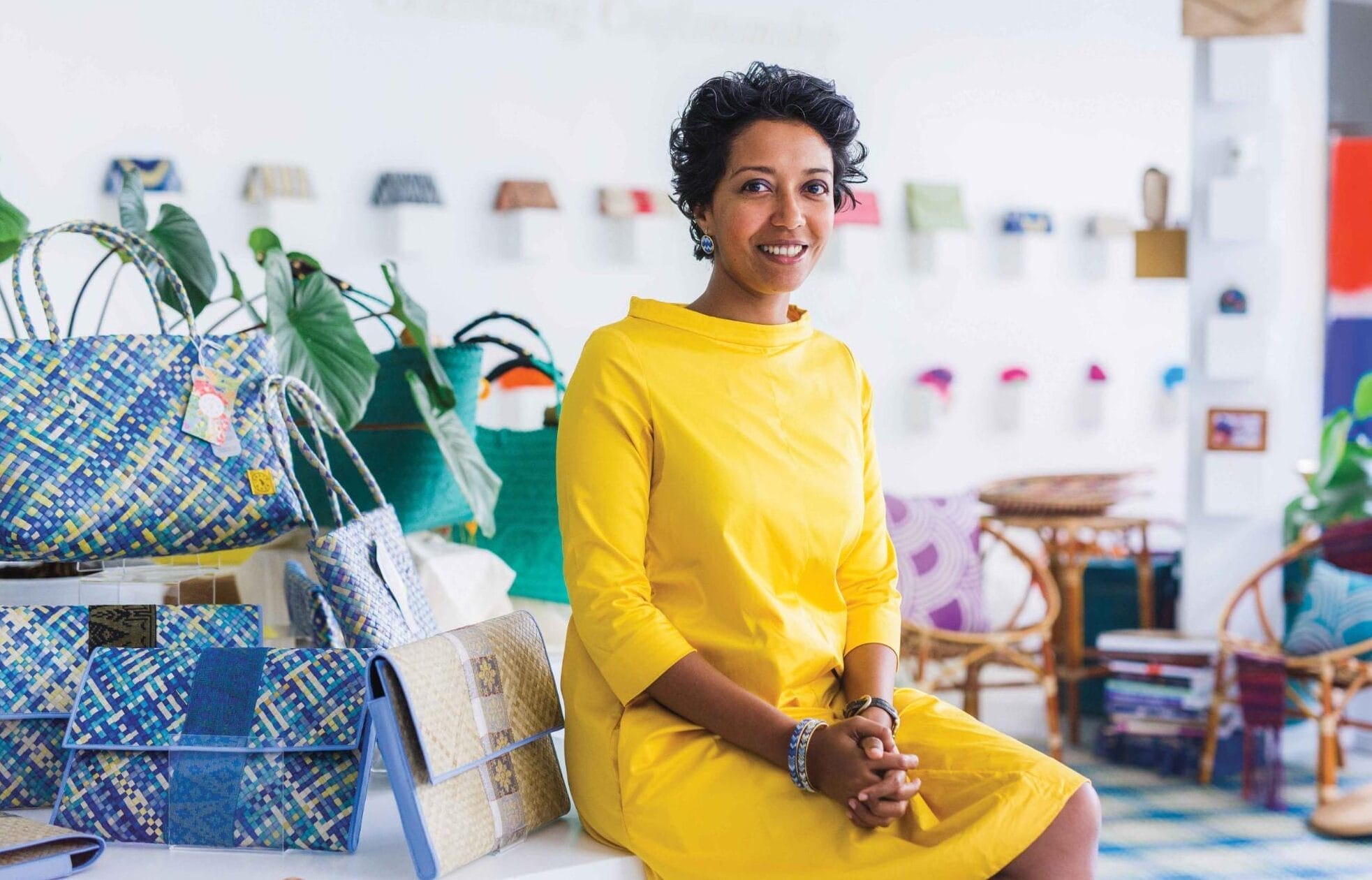 A woman sits in a yellow dress next to a few handbags.