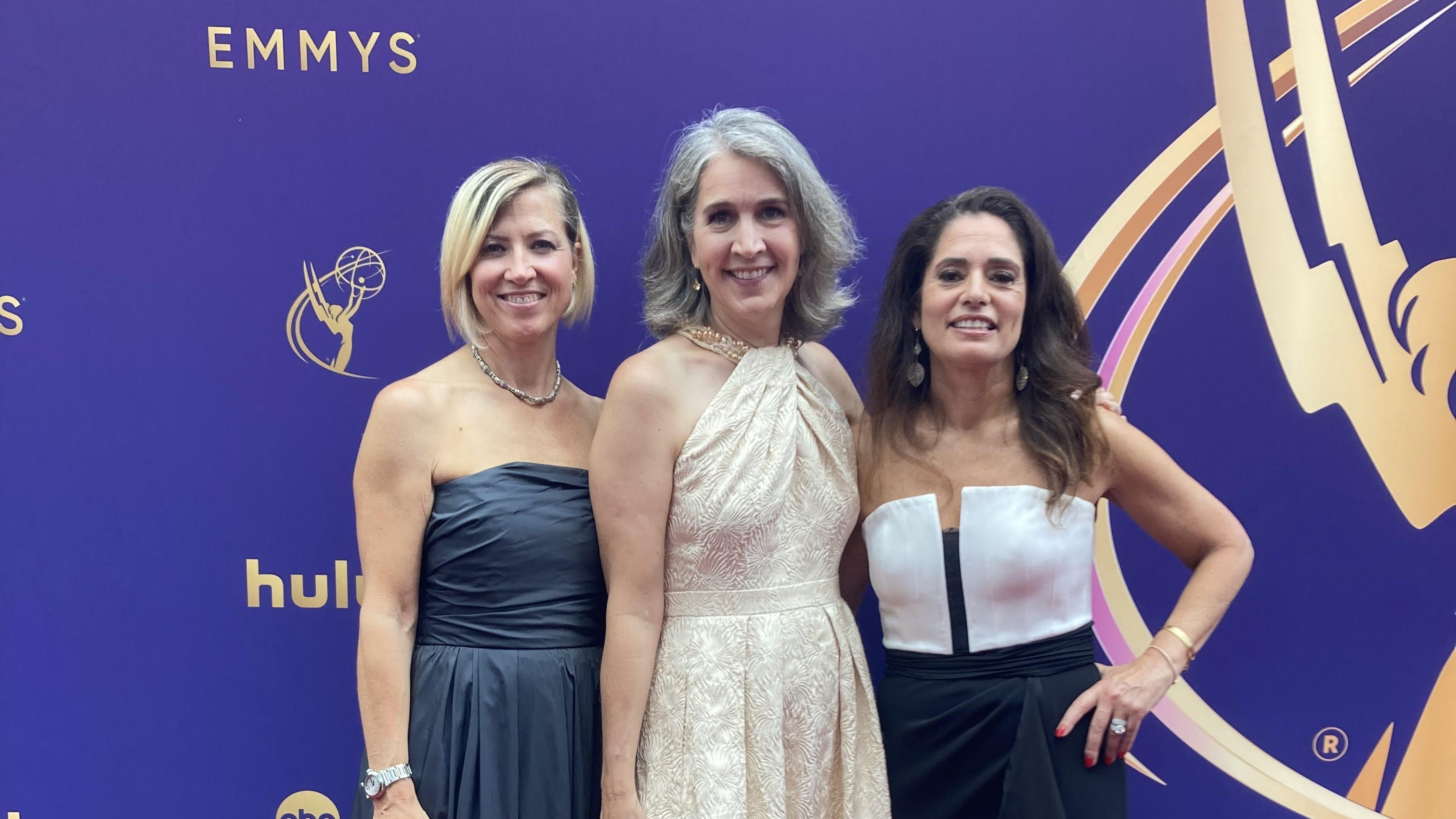 Three alumnae in elegant dresses stand in front of an Emmys backdrop.