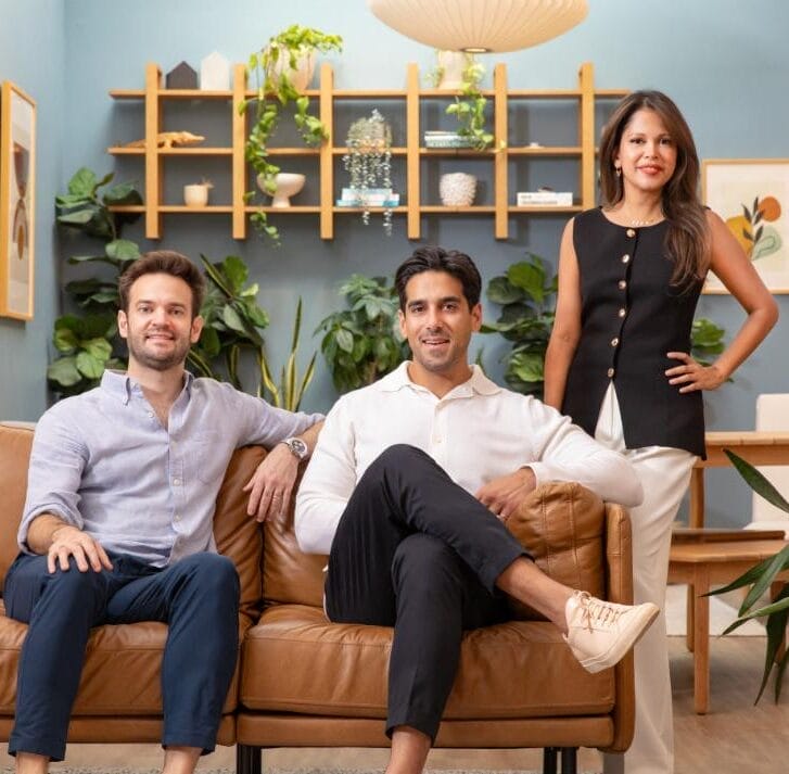 Two men sit on a couch with a woman posed behind them with her hand on her hip.