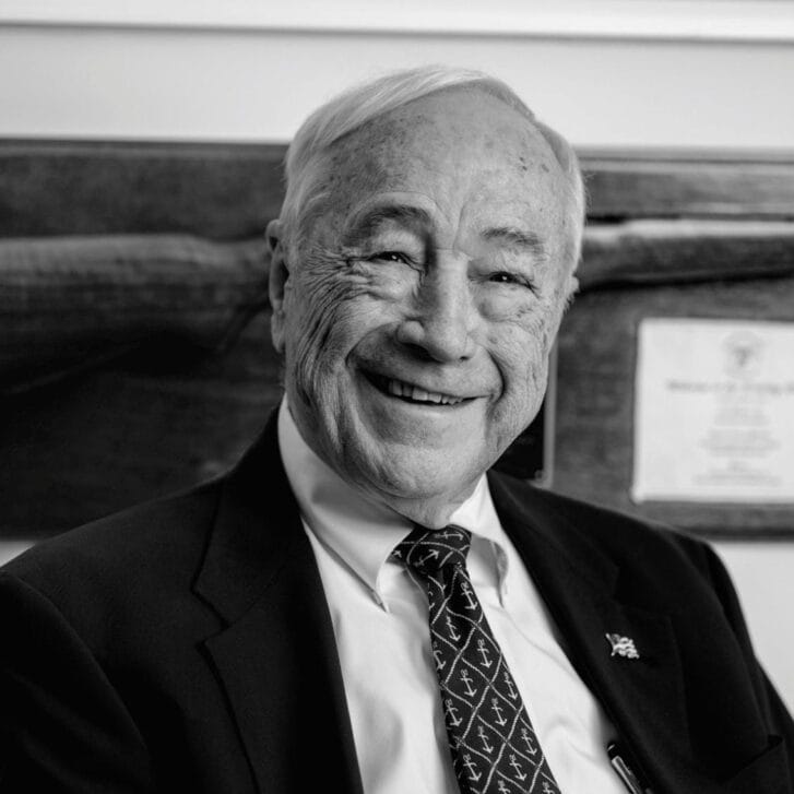 Man in a black suit jacket, white button-down shirt, and a tie with anchors patterned on it.