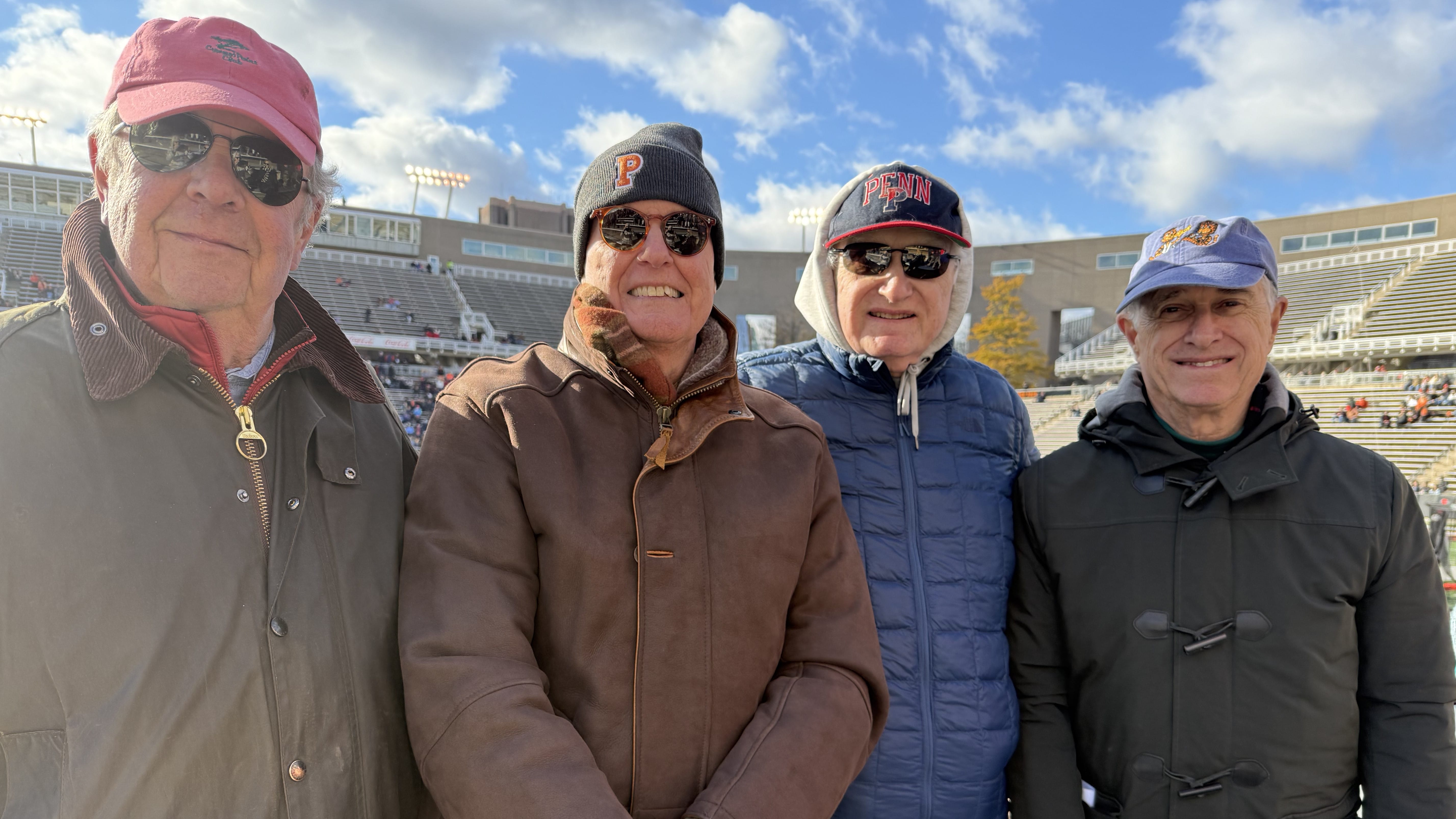 Four men in winter jackets and caps pose for a photo in a stadium.