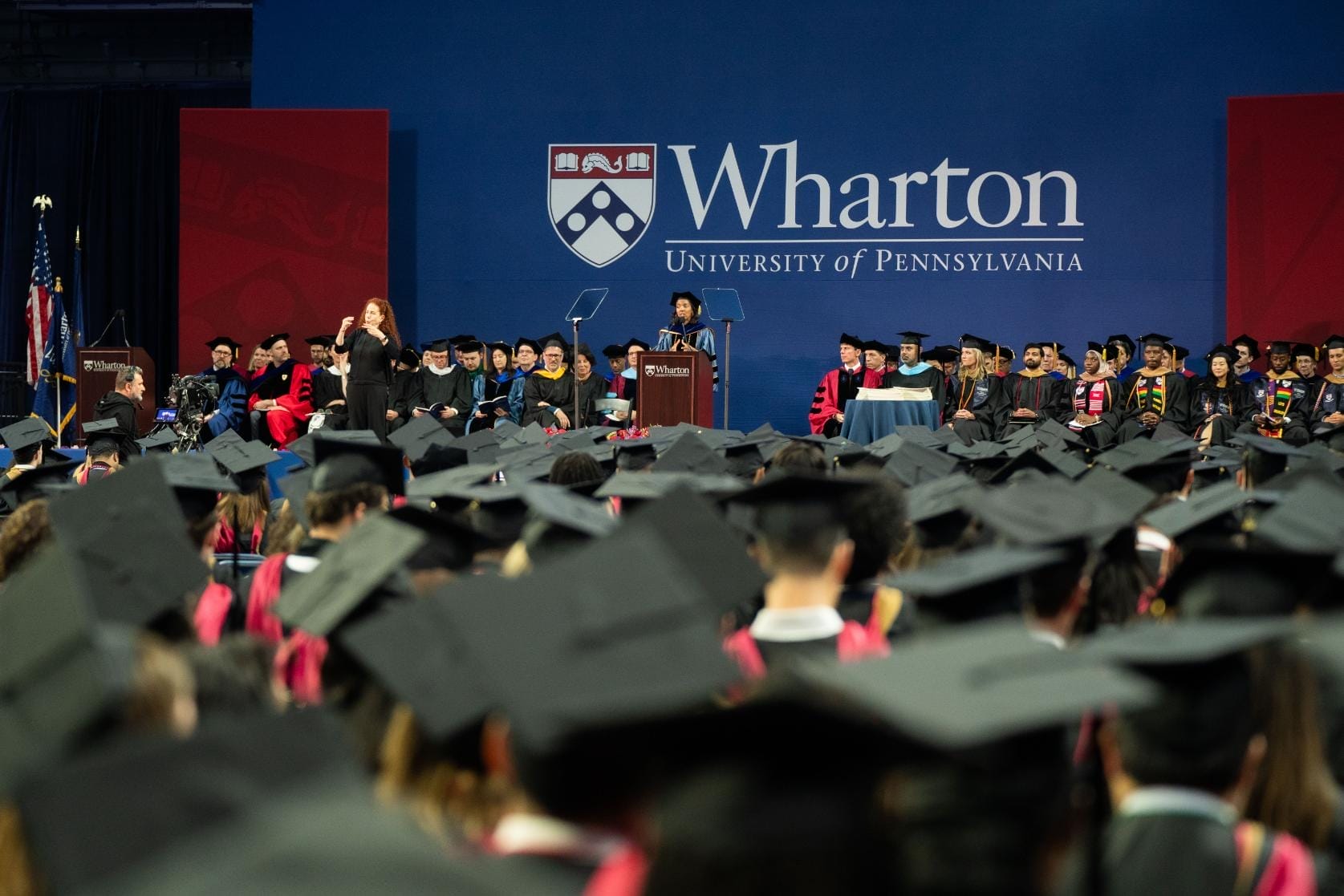 Students wearing black caps and gowns seated in front of a graduation stage.