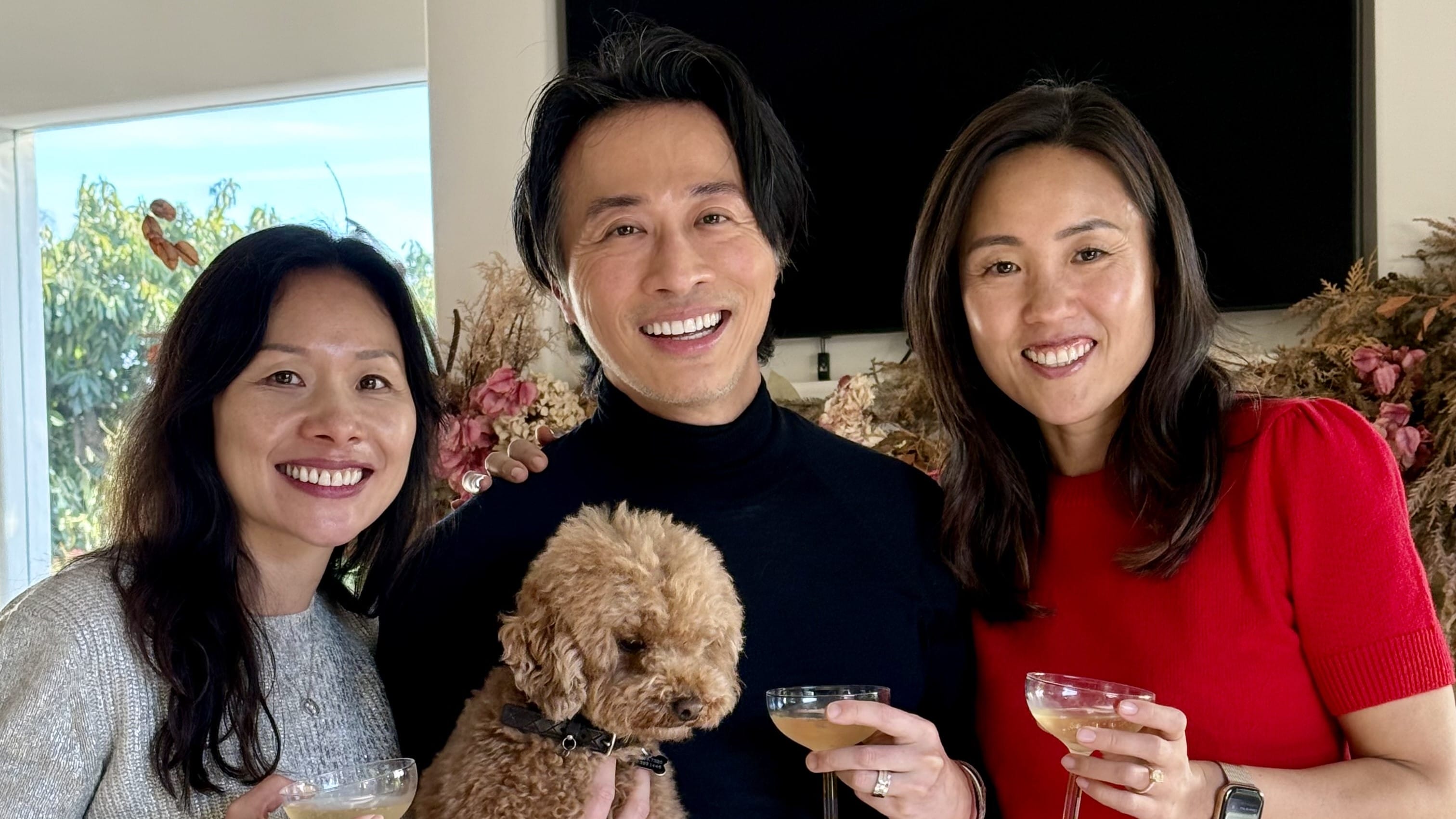 Three people in sweaters pose together holding drinks and a dog.