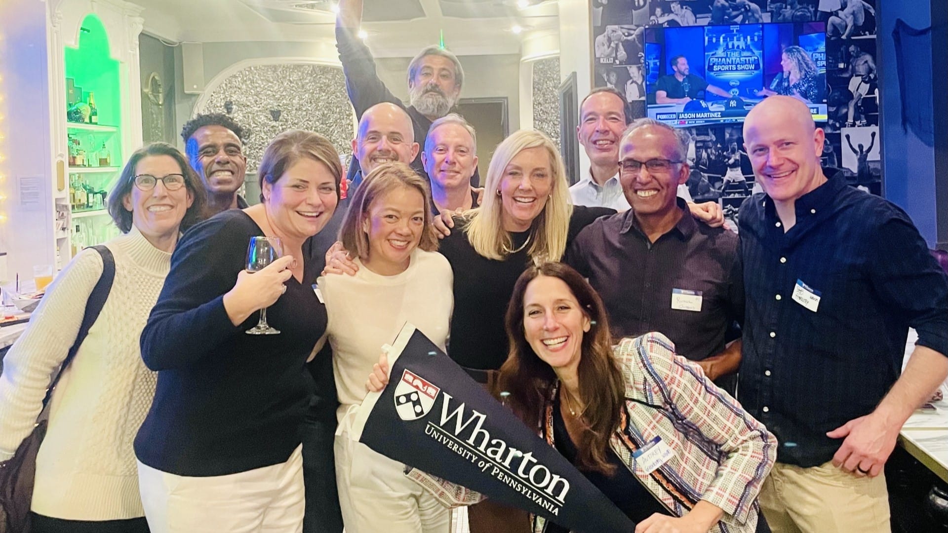 A dozen alumni in nice attire gather for a photo behind a Wharton flag one alumna is holding up.