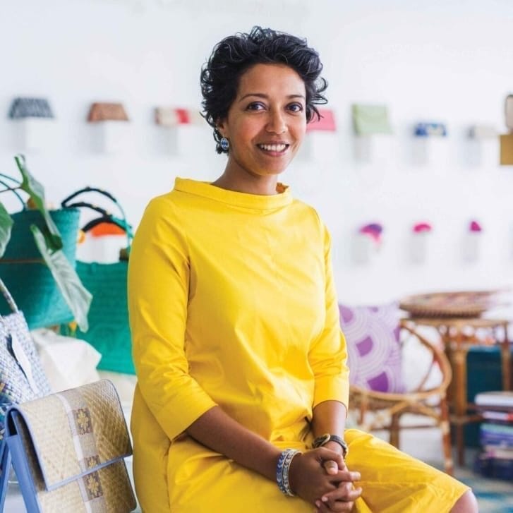 A woman sits in a yellow dress next to a few handbags.