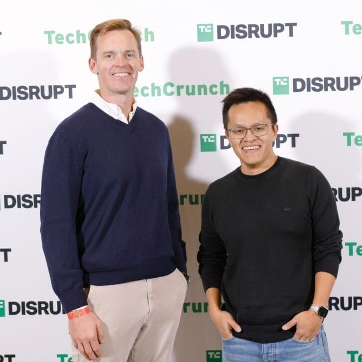 Two men stand in long sleeve shirts posed in front of a photo background that says TechCrunch Disrupt.