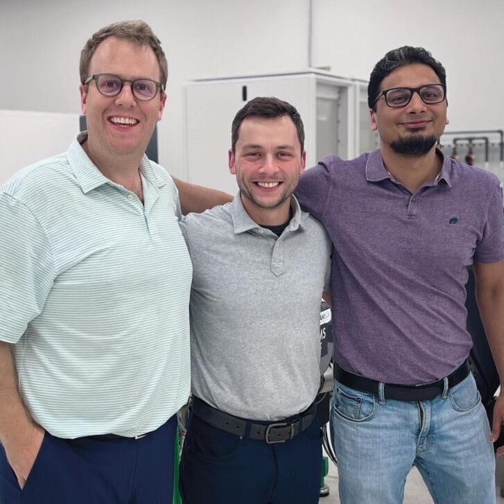 Three men stand shoulder to shoulder wearing polos in different colors in a lab-like setting.