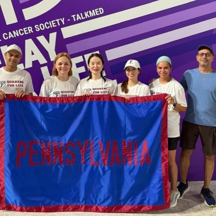 A group of Wharton alumni stands in front of a purple Relay for Life backdrop holding a flag that says 