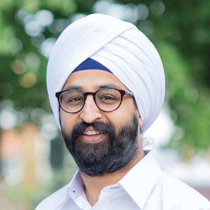 A portrait of a man wearing a dastār (turban), black-rimmed glasses, and a white button-down shirt.